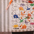 HeyDoodle DinoRoar silicone mat with dinosaurs, volcanoes, and trees, partly colored on a striped tablecloth for creative play.
