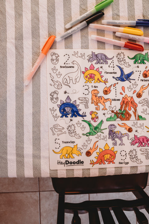 HeyDoodle DinoRoar silicone mat with dinosaurs, volcanoes, and trees, partly colored on a striped tablecloth for creative play.