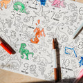 A reusable silicone HeyDoodle DinoRoar mat on tiles, partly colored with green, blue, pink, and orange; features dinos and volcano.