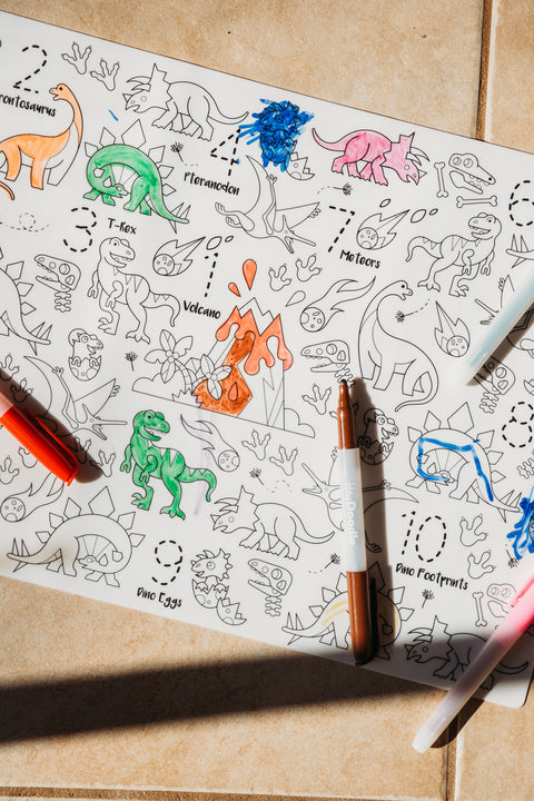 A reusable silicone HeyDoodle DinoRoar mat on tiles, partly colored with green, blue, pink, and orange; features dinos and volcano.