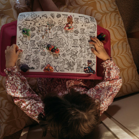 A child colours vibrant doodles and numbers on a reusable silicone HeyDoodle mat, using markers amid toys and a cozy cushion.