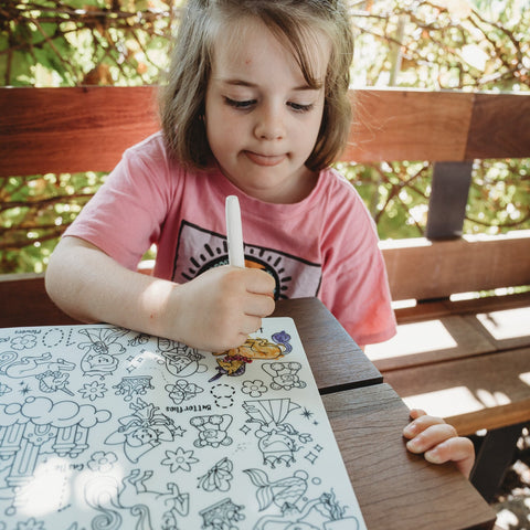 A young girl in a pink shirt sits on a wooden bench outdoors, coloring numbers on a HeyDoodle silicone mat filled with playful designs.