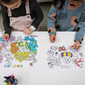 Two young children color vibrant native flowers on the reusable HeyDoodle silicone mat, markers and their cup ready for more fun.