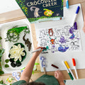 Two kids colour on the HeyDoodle Into The Wild reusable silicone mat at a table with toy animals, markers, and a book.