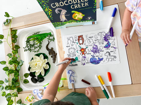 Two kids colour on the HeyDoodle Into The Wild reusable silicone mat at a table with toy animals, markers, and a book.