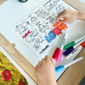 A child colours a silicone HeyDoodle Toot Toot Honk mat with transport designs, using markers. Extra pens, yellow kinetic sand tray and markers scattered.