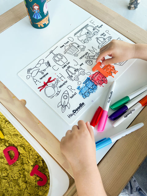 A child colours a silicone HeyDoodle Toot Toot Honk mat with transport designs, using markers. Extra pens, yellow kinetic sand tray and markers scattered.