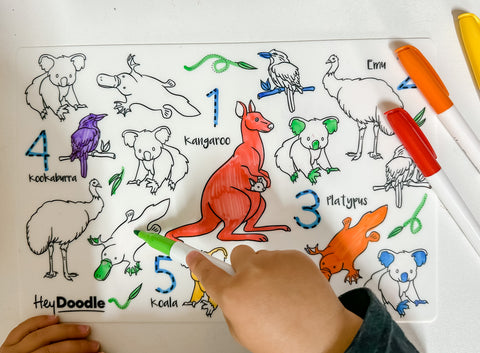 A child colours a reusable silicone HeyDoodle mat of Aussie animals—kangaroo, emu, platypus, koala, kookaburra; two markers nearby.