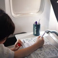 A child colours on a silicone HeyDoodle Toot Toot Honk Mat with markers on a plane tray; pens sit in a branded cup nearby.