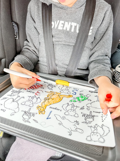 A child in a car seat colours animal designs on the reusable HeyDoodle silicone mat with red and yellow markers on a baking tray.