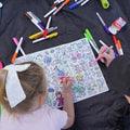 Two kids color bright cartoon designs on a black silicone HeyDoodle mat; one with a white bow practices numbers as they create.