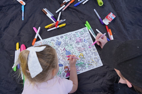 Two kids color bright cartoon designs on a black silicone HeyDoodle mat; one with a white bow practices numbers as they create.