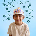 A smiling child wears a Royal Crown - Wearable & Reusable Colouring Crown featuring their own custom colouring, surrounded by floating star and confetti illustrations.