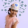 A young girl smiles while wearing a HeyDoodle Australia Tiara Hearts, an adjustable silicone crown featuring hand-drawn hearts and swirls designed to be coloured and reused.