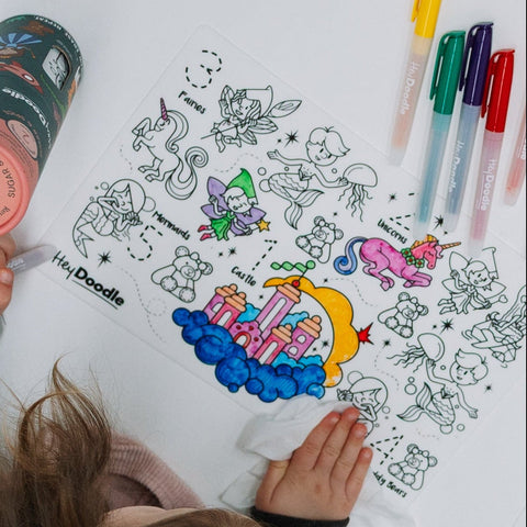 A child colours on a reusable silicone HeyDoodle mat with fairies, unicorns, castles and stars; markers nearby for endless fun.