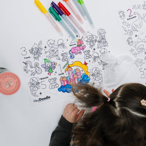 A young child with brown pigtails using colourful silicone HeyDoodle mat decorated with fairies, unicorns, and a castle.