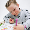 A young child with braided hair colors a silicone HeyDoodle mat with a pink washable crayon pen, markers on a white table nearby.