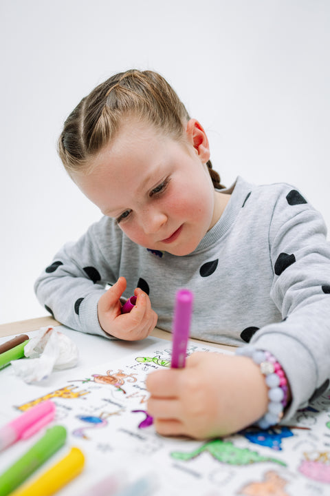 A young child with braided hair colors a silicone HeyDoodle mat with a pink washable crayon pen, markers on a white table nearby.