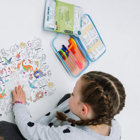 A child with braided hair colors dinosaurs and practices numbers on a silicone HeyDoodle DinoRoar mat, using markers from a blue case, all placed on a white surface for creative colouring fun.
