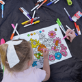 A child with a white bow and an adult color a reusable silicone HeyDoodle colouring mat, surrounded by scattered vibrant markers.