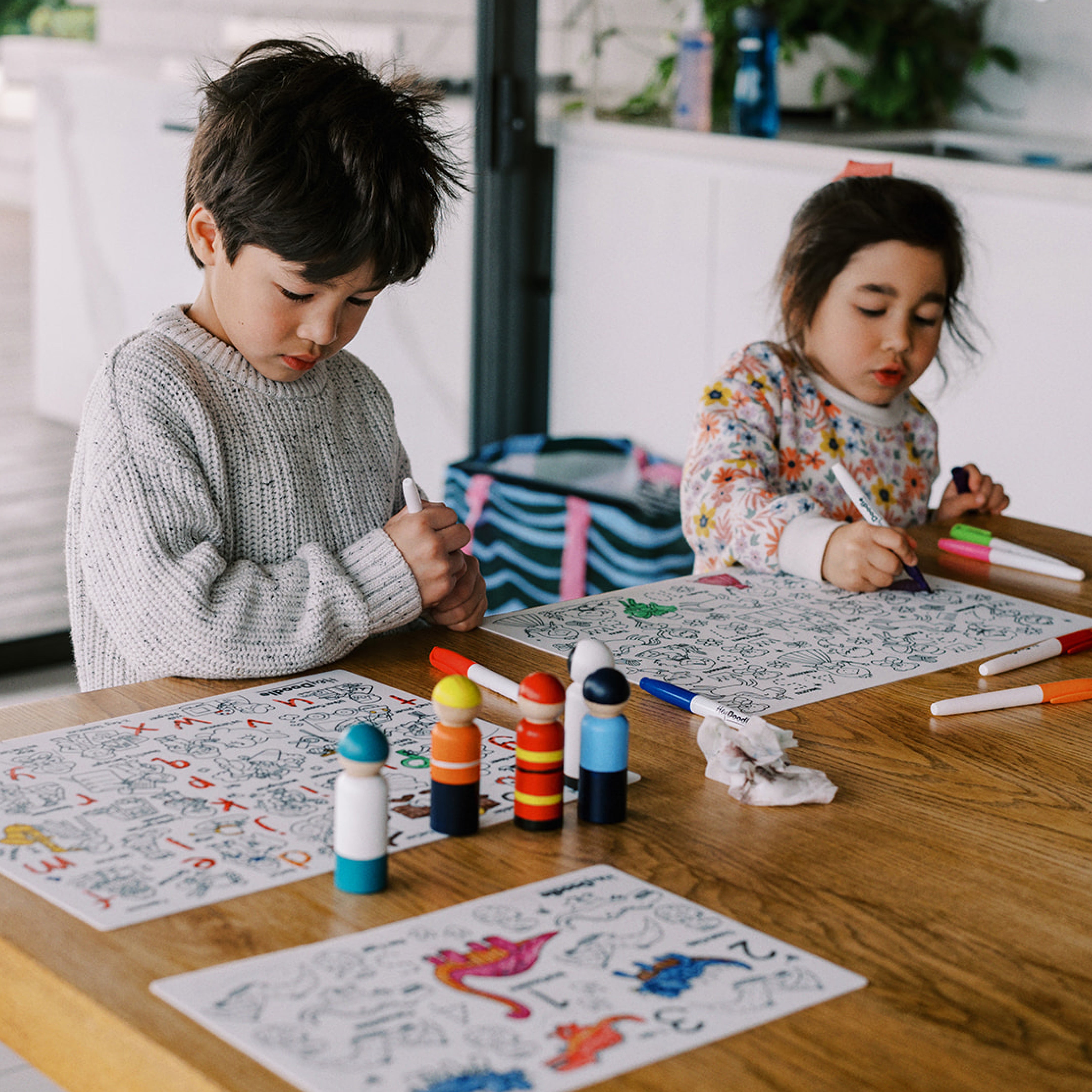 Two children color on vibrant silicone activity mats using colorful markers and daubers at a wooden table in a bright kitchen.