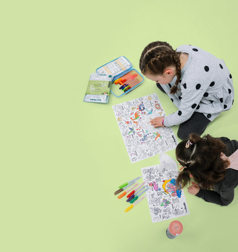 Two kids color a vibrant reusable silicone mat covered in playful designs with markers. Art tools and a drink sit nearby, seen from above.