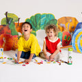 Two young children laugh as they colour on HeyDoodle’s reusable silicone activity mat with vibrant crayons. Surrounding them are bright, cut-out silicone illustrations of fruit, leaves, and a caterpillar inspired by The Very Hungry Caterpillar. The HeyDoodle Australia The Very Hungry Caterpillar™ Mini Colouring & Doodle Mat and other colourful silicone art supplies are scattered nearby, offering a playful and portable way for kids to create again and again wherever they go.