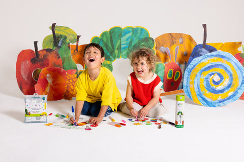 Two young children laugh as they colour on HeyDoodle’s reusable silicone activity mat with vibrant crayons. Surrounding them are bright, cut-out silicone illustrations of fruit, leaves, and a caterpillar inspired by The Very Hungry Caterpillar. The HeyDoodle Australia The Very Hungry Caterpillar™ Mini Colouring & Doodle Mat and other colourful silicone art supplies are scattered nearby, offering a playful and portable way for kids to create again and again wherever they go.