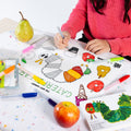 A child in a pink sweater colors on The Very Hungry Caterpillar™ Classic Colouring & Doodle Mat by HeyDoodle Australia at a small table, surrounded by colorful markers, pieces of fruit, a water bottle, and an open book. The vibrant silicone mat features themed designs to spark creativity, providing a mess-free and reusable activity that encourages imaginative coloring again and again.