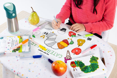 A child in a pink sweater colors on The Very Hungry Caterpillar™ Classic Colouring & Doodle Mat by HeyDoodle Australia at a small table, surrounded by colorful markers, pieces of fruit, a water bottle, and an open book. The vibrant silicone mat features themed designs to spark creativity, providing a mess-free and reusable activity that encourages imaginative coloring again and again.