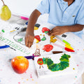 A young boy erases a marker drawing from the HeyDoodle Australia The Very Hungry Caterpillar™ Classic Colouring & Doodle Mat, a colourful silicone activity mat, on a white table scattered with other vibrant silicone colouring mats, fruit, markers, and a copy of The Very Hungry Caterpillar book—creating an inviting screen-free creative space for children to colour and doodle again and again.