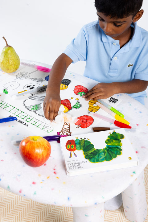 A young boy erases a marker drawing from the HeyDoodle Australia The Very Hungry Caterpillar™ Classic Colouring & Doodle Mat, a colourful silicone activity mat, on a white table scattered with other vibrant silicone colouring mats, fruit, markers, and a copy of The Very Hungry Caterpillar book—creating an inviting screen-free creative space for children to colour and doodle again and again.
