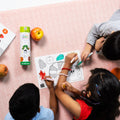 Three children are colouring together on a pink patterned surface, using the HeyDoodle Australia The Very Hungry Caterpillar™ Mini Colouring & Doodle Mat. The silicone mat features vibrant designs from the classic story and is surrounded by apples, colourful markers, and a juice box. Fully engaged in their creative activity, the kids enjoy this reusable, travel-friendly silicone mat that keeps them entertained and inspired wherever they go.
