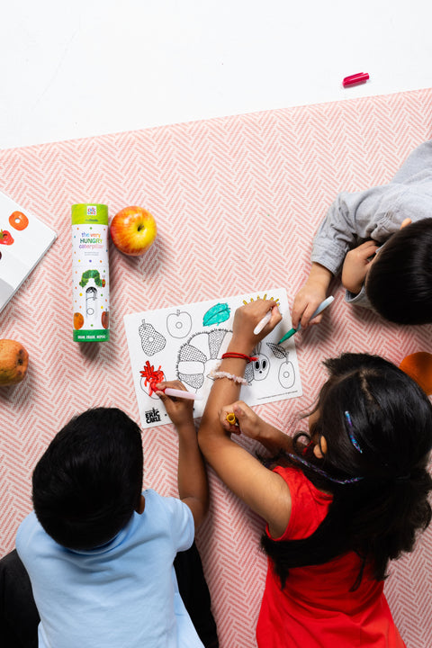 Three children are colouring together on a pink patterned surface, using the HeyDoodle Australia The Very Hungry Caterpillar™ Mini Colouring & Doodle Mat. The silicone mat features vibrant designs from the classic story and is surrounded by apples, colourful markers, and a juice box. Fully engaged in their creative activity, the kids enjoy this reusable, travel-friendly silicone mat that keeps them entertained and inspired wherever they go.