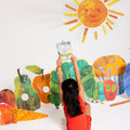 A child dressed in red sits on the floor, holding up a book. Around them, vibrant silicone cutouts of fruits, a caterpillar, and a sun adorn the white background—setting the scene for engaging, screen-free play. This cheerful setup perfectly showcases HeyDoodle’s The Very Hungry Caterpillar™ Classic Colouring & Doodle Mat: a reusable silicone activity mat designed to spark creativity and offer endless opportunities for colourful doodling and imaginative fun.