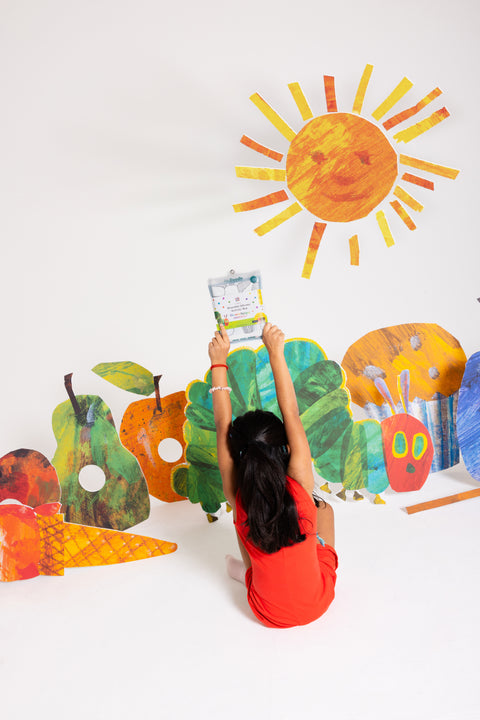 A child dressed in red sits on the floor, holding up a book. Around them, vibrant silicone cutouts of fruits, a caterpillar, and a sun adorn the white background—setting the scene for engaging, screen-free play. This cheerful setup perfectly showcases HeyDoodle’s The Very Hungry Caterpillar™ Classic Colouring & Doodle Mat: a reusable silicone activity mat designed to spark creativity and offer endless opportunities for colourful doodling and imaginative fun.