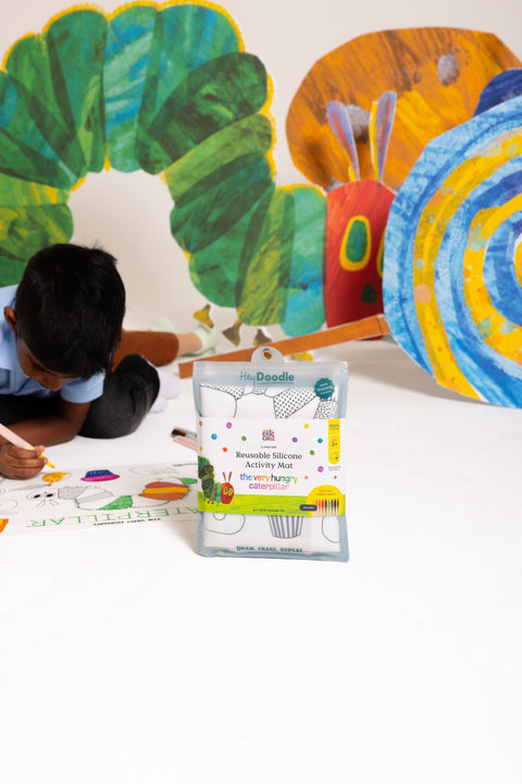 A child engages with vibrant The Very Hungry Caterpillar-themed silicone mats, joyfully colouring and enjoying a screen-free creative activity. In the foreground, the HeyDoodle Australia The Very Hungry Caterpillar™ Classic Colouring & Doodle Mat package stands upright, highlighting its colourful and playful reusable silicone mat design perfect for imaginative play.