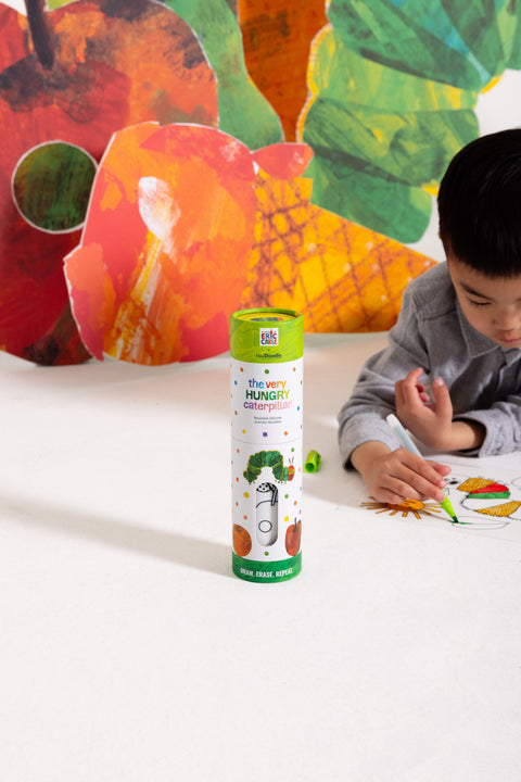 A child sits on the floor using colourful markers to decorate a reusable silicone HeyDoodle Australia The Very Hungry Caterpillar™ Mini Colouring & Doodle Mat, surrounded by a lively caterpillar-themed scene filled with bright colours and playful patterns.