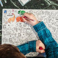 A child colours an orange T-Rex and green plants on a HeyDoodle silicone mat featuring dinosaurs and creative, reusable scenes.