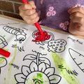 A child in a purple shirt with pink flowers colours a strawberry on a silicone HeyDoodle mat using washable crayons to fill outlines and colour words like flower, caterpillar, and carrot.