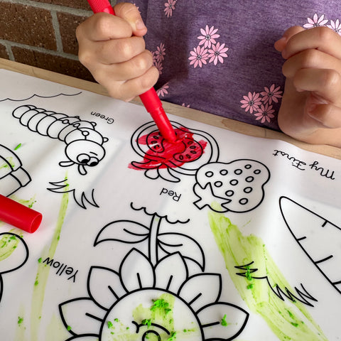 A child in a purple shirt with pink flowers colours a strawberry on a silicone HeyDoodle mat using washable crayons to fill outlines and colour words like flower, caterpillar, and carrot.