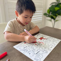 A young boy colours on a HeyDoodle  DinoRoar silicone mat with a red marker; other markers laying around and a green plant behind.