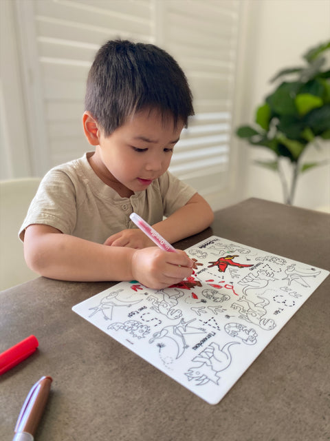 A young boy colours on a HeyDoodle  DinoRoar silicone mat with a red marker; other markers laying around and a green plant behind.
