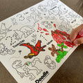 A child's hand colors a silicone DinoRoar Mini Mat by HeyDoodle, using a red marker on outlined dinosaurs, volcanoes, and numbers.