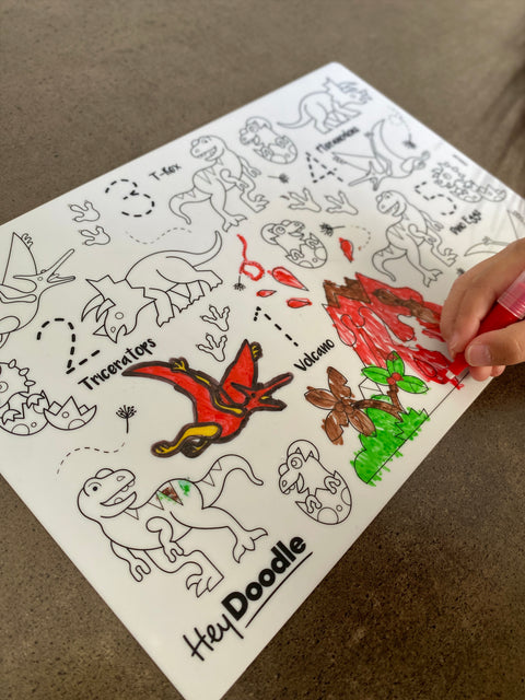 A child's hand colors a silicone DinoRoar Mini Mat by HeyDoodle, using a red marker on outlined dinosaurs, volcanoes, and numbers.