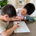 Two young boys sit with a HeyDoodle DinoRoar silicone mat—one colors with green while the other, holding the tube, watches intently.