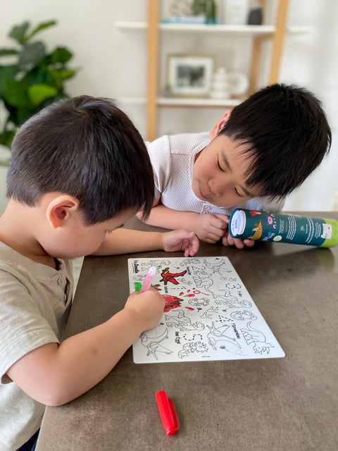 Two young boys sit with a HeyDoodle DinoRoar silicone mat—one colors with green while the other, holding the tube, watches intently.