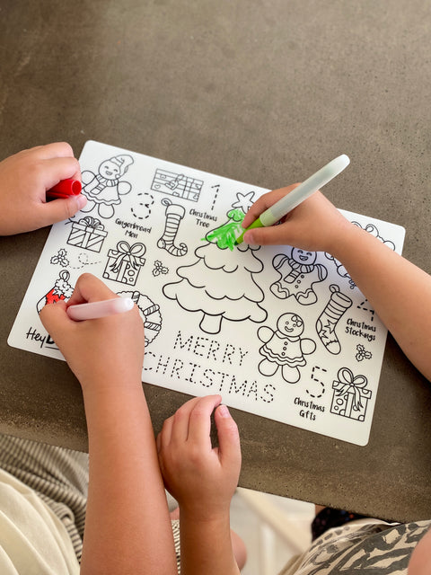 Two kids colour a reusable HeyDoodle Jingle Bells silicone mat with festive trees, gingerbread, gifts, stockings, and numbers.