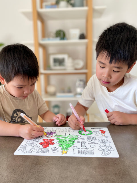 Two boys color the reusable HeyDoodle silicone mat, featuring a festive tree and holiday designs. 