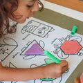 A young child shapes colored dough on a reusable silicone HeyDoodle mat, matching a triangle outline among playful labeled animals.